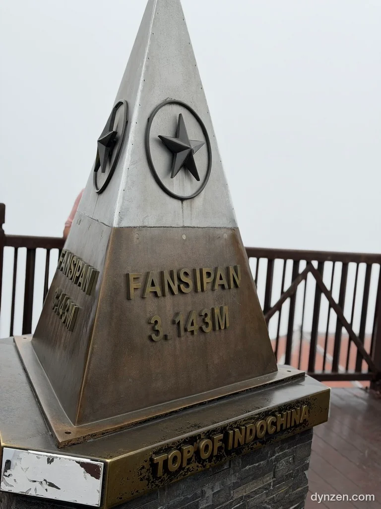 삼각뿔 모양의 정상 표지석, ‘FANSIPAN 3.143M’과 ‘TOP OF INDOCHINA’가 새겨져 있고 뒤는 온통 흰 안개