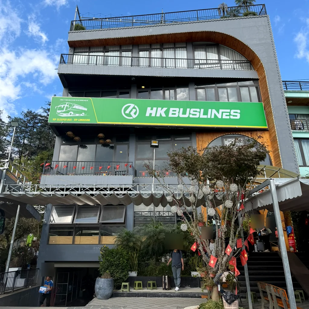 HK BUSLINES 초록 간판이 걸린 사파 오피스 건물, 파란 하늘 아래 계단 앞으로 여행객이 오간다
