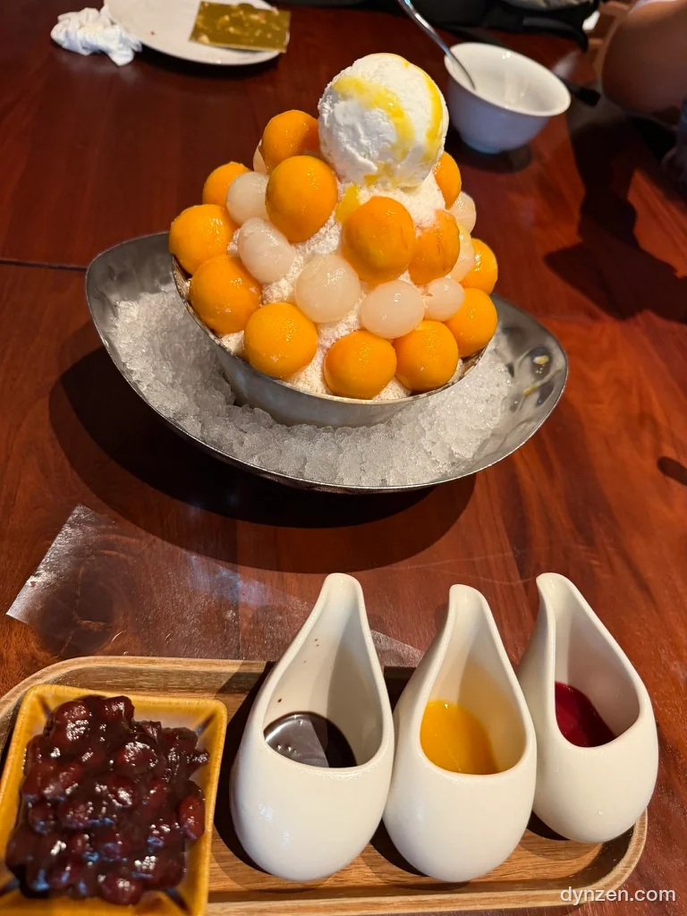 철제 그릇에 쌓아 올린 동그란 생망고 조각과 아이스크림을 올린 망고 빙수, 앞에 팥과 시럽이 담긴 작은 그릇들