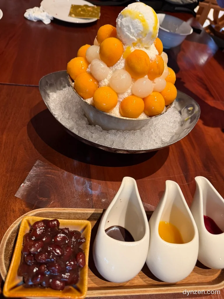 철제 그릇에 담은 망고빙수, 동그랗게 떠낸 생망고와 리치를 쌓고 꼭대기에 아이스크림을 올렸다, 그릇 둘레에 얼음을 깔았고 앞쪽에 팥과 소스 세 가지가 놓여 있다